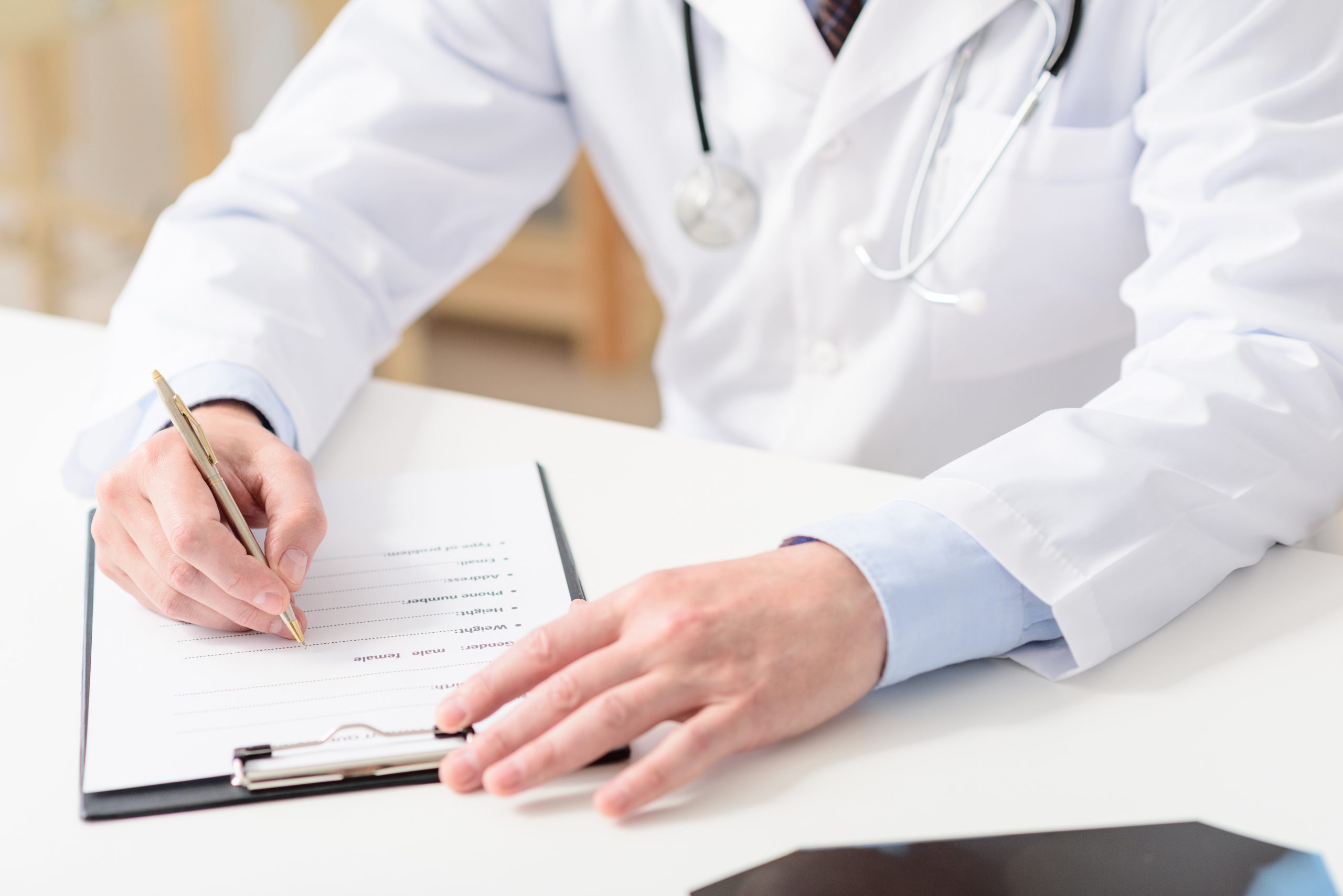 Smiling pediatrician sitting at table Accessing medical records. Close up of doctor writing on his clipboard, sitting in his office